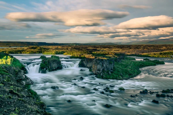 空に白い雲と複数の小さな岩の間を流れる水の日の出の間にアイスランドの泥炭の滝の長い露出の写真 — ストック写真