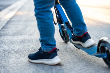 Kaldırımda mavi tekmeli scooter süren kot ve spor ayakkabılı çocuk bacakları, kentsel gençlerin ulaşımını ve aktif yaşam tarzını gösteriyor