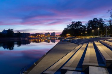 Polonya şehir manzarasında yansımalar ve parlayan ışıklarla nehir kenarındaki gezinti alanı üzerinde renkli bir akşam alacakaranlığı. Şehir manzarası için uygun.