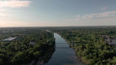 Bereketli yeşil nehir kenarı parkı ve gün batımında yaya köprüsü manzarası, huzurlu kentsel doğa görüntüleri.