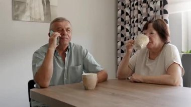 Yaşlı çift evde kahve içip sohbet ederken, yaşlı adam rahat bir atmosferle akıllı telefondan konuşuyor.
