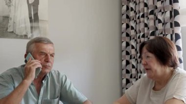 Yaşlı adam evde ciddi bir telefon görüşmesi yapıyor, yaşlı çift mutfak masasında düşünceli bir anı paylaşıyor.