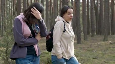 Su şişesi ve tasmalarla ormanda yürüyen iki kadın, sohbet ve fiziksel aktivite içindeler.