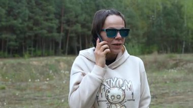 Güneş gözlüklü ve kapüşonlu bir kadın ormanda cep telefonuyla konuşurken gülümsüyor. Kamera onun etrafında dönüyor.