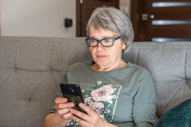 Polonya 'da gözlüklü yaşlı bir kadın akıllı telefon kullanarak koltukta oturuyor.