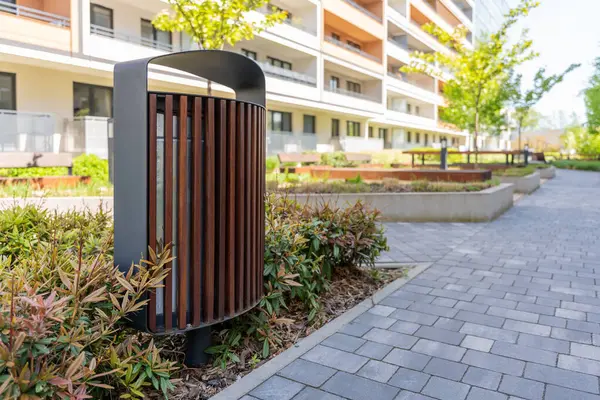Modern apartman binasının bahçesinde siyah çerçeveli ahşap çöp tenekesi, kentsel mobilya tasarımını gösteriyor.