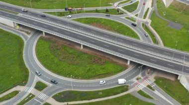 İnsansız hava aracı görüntüleri, şehir trafik altyapısını gösteren, yeşilliklerle çevrili, modern çok şeritli bir otoyolda ve kavşakta gezinen araçları gösteriyor.