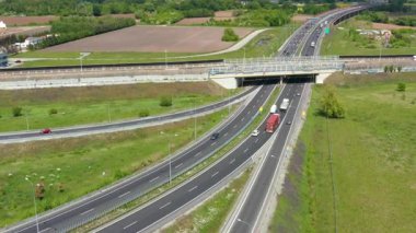 Görünür üst geçitli bir otoyol kavşağının drone görüntüleri, hareket halindeki arabalar ve kamyonlar, yeşil alanlarla çevrili ve açık gökyüzünün altında seyrek altyapı