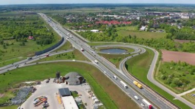Yeşil tarlalar ve yerleşim bölgeleriyle çevrili su tutma göletinin etrafındaki rampalarda gezinen araçlarla yonca yaprağı kavşağının insansız hava aracı görüntüleri.