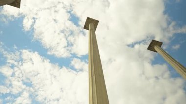 Eski mimariyi andıran yüksek klasik beton sütunların alçak açılı perspektifi, parçalı bulutlu mavi gökyüzüne bakarak, ölçeği, tarihi ve tasarımı vurguluyor. Kamera yaklaşımı ve sütun etrafında döndürme.