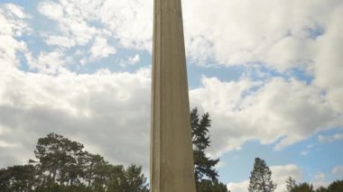 Etrafı ağaçlarla çevrili parçalı bulutlu bir gökyüzüne yükselen beton sütunların zemin manzarası, halka açık bir alanda heybetli ve sakin bir atmosfer hissi uyandırıyor. Kamera yukarı eğ.