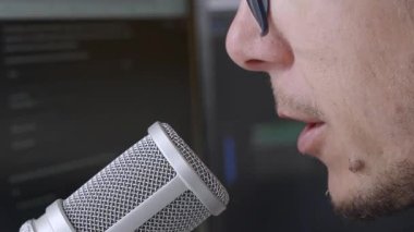 Arka planda bilgisayar monitörü olan profesyonel bir mikrofona konuşan bir adamın yakın plan çekimi, podcast ya da seslendirme gibi modern içerik oluşumunu gösteriyor.