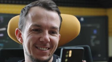 Modern bir çalışma alanında oturan neşeli bir yetişkin adam akıllı telefonuna bakarken gülümsüyor. Ofiste birden fazla monitör ve şık bir sandalye var. Teknoloji meraklısı olduğunu gösteriyor..