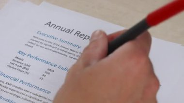 Yıllık basılı mali raporda anahtar verileri işaretlemek için kırmızı kalem kullanan birinin elinin yakın plan görüntüsü. Görüntüler iş incelemesi ve analitik çalışmaları gösteriyor.