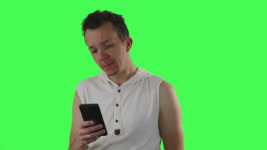 Kolsuz beyaz kapüşonlu genç adam şaşkınlık ve şaşkınlık da dahil olmak üzere duygusal ifadeleri olan akıllı telefona bakıyor, kompozitasyon ve hikaye anlatma için yeşil ekran arka planında görüntüleniyor.