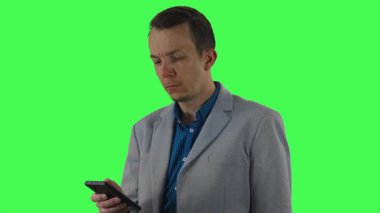 Gri ceketli orta yaşlı bir adam, iş iletişiminde veya teknoloji temalarında çok yönlü kullanım için yeşil krom anahtar arka planına karşı çekilen akıllı telefonuyla iletişim kurarken odaklanmış ve endişeli görünüyor..