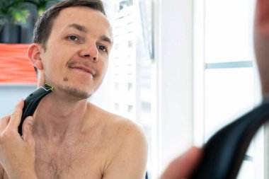 Ayna yansımasında elektrikli budayıcıyla tımarcılık yapan genç bir adam. Parlak banyo içinde odaklanmış bir ifade.