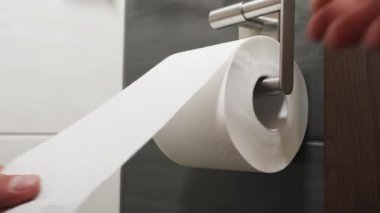 Eller, koyu fayanslı banyo duvarına monte edilmiş, beyaz tuvalet kağıdını dikkatlice açar. Temizlik, hijyen uygulamaları ve kişisel bakım alışkanlıkları sergiler.