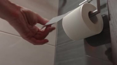 Bir adamın eli tuvalet kağıdı rulosundan bir çarşaf çıkarır ve banyonun içindeki fayanslı bir duvara monte edilmiş metal bir tutacağa asılır. Görüntüler basit bir hijyen rutinini gösteriyor..