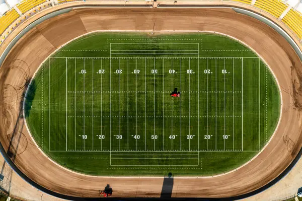 Amerikan futbol sahası ve yeşil alanda çalışan iki kırmızı traktörlü oval spor stadyumunun yukarıdan aşağı görüntüsü.