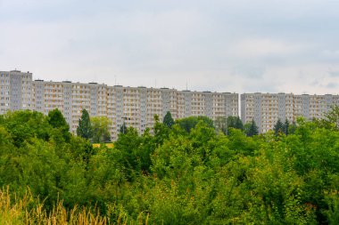 Bulutlu gökyüzünün altındaki yemyeşil ağaçların arkasındaki aynı gri apartman blokları. Komünizm sonrası konut kavramı.