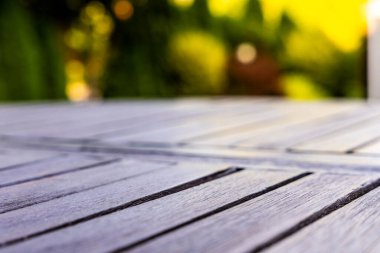 Arka planda bulanık yaz yeşilliği olan ahşap bahçe terası tahtalarının yakın görüntüsü, öğleden sonra ışığı altında yakalanmış, ürün yerleştirme ve dış mekan kavramları için kullanışlı.