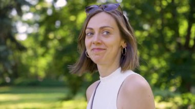 Güneşli bir yaz gününde yeşil bir parkta huzurlu bir yürüyüş yapan kısa kahverengi saçlı ve güneş gözlüklü genç bir kadının yavaş çekimde çekilmiş portresi sakinliğini ve doğal güzelliğini gösteriyor..
