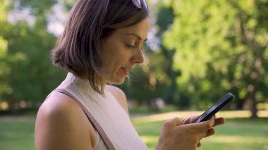 Yavaş çekim videosu, akıllı telefonlu dokunmatik ekranı ile meşgul olan genç bir kadının, güneş ışığında yeşil bir parkta durarak doğadaki gevşek dijital iletişimi ön plana çıkardığını gösteriyor..