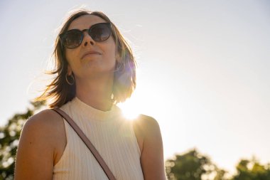Güneş gözlüklü, kendine güveni tam bir kadın. Yeşil parkta akşam güneşinin altında duruyor. Güneş ışığı, huzurlu yaz havasının altını çiziyor.