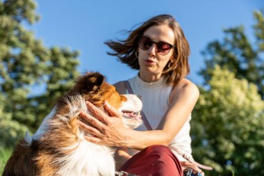 Neşeli bir kadının güneşli bir parkta köpekle bağ kurması, günlük giysiler ve doğal etkileşimler.