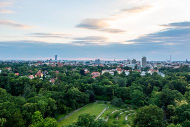 Bereketli yeşil parkın ve bulutlu günbatımı gökyüzünün altındaki uzak şehir manzarasının hava manzarası.