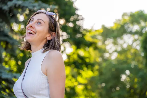 Güneşli parkta gülümseyen ve bakan mutlu genç bir kadın. Arka planda bulanık yeşil ağaçlar, neşeli yaz günü açık hava yaşam tarzı anı.