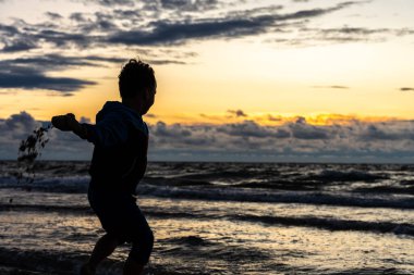 Gün batımında deniz kenarında oynayan çocuk, sahile kum, siluet, enerji ve çocukluk özgürlüğü fırlatıyor.