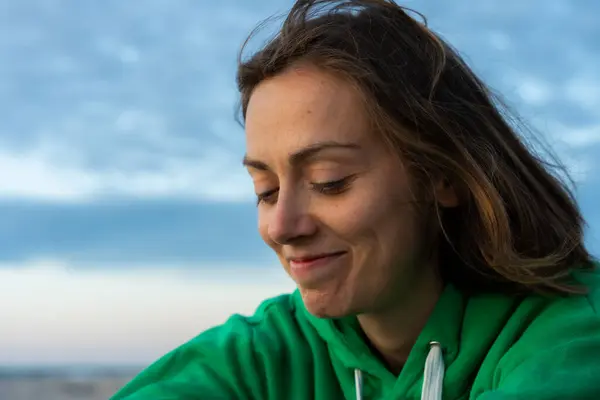 Yeşil kapüşonlu gülümseyen kadının yakın plan portresi aşağıya bakıyor, kumsalda alacakaranlıkta yakalanmış.