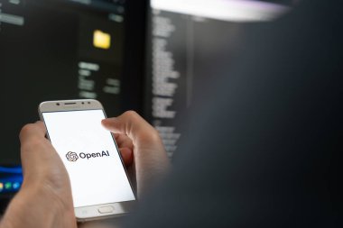 OpenAI logosuyla el ele tutuşan akıllı telefon, arka planda bilgisayar kodu olan karanlık bir ortamda ekranda.