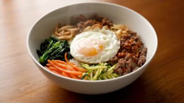 Lezzetli bir kase bibibimbap, kızarmış yumurta ve taze sebzelerle ahşap bir masada hazır bekliyor.