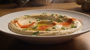 Kırmızı biber ve maydanozlu, tahta bir yüzey masasının üstünde beyaz bir kasede yakın plan bir humus fotoğrafı.