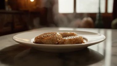 Susam tohumlu buharda pişen et parçaları mermer bir masa yüzeyinde beyaz bir tabakta sunuluyor.