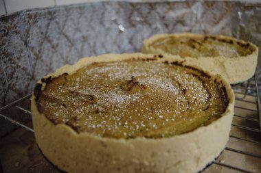 Two freshly baked pumpkin pies cooling on a wire rack, with a dusting of powdered sugar on top. The background features a reflective, patterned surface.