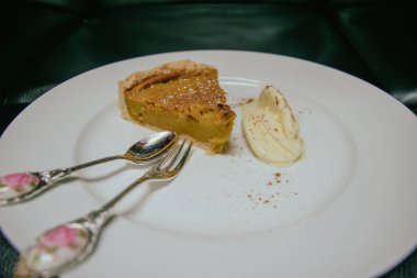 A slice of pumpkin pie on a white plate with a dollop of whipped cream and two ornate silver utensils. The pie is sprinkled with cinnamon.