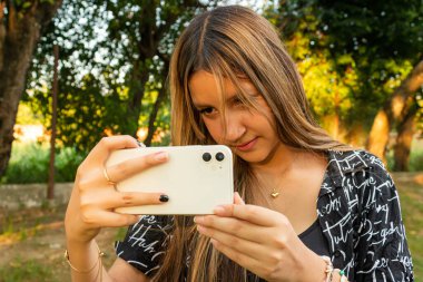 Beyaz akıllı telefonuyla yeşil alanda fotoğraf çeken Latin bir genç kadın.