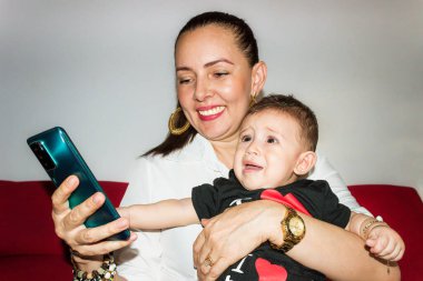 Küçük oğlu kollarında ağlarken neşeli Latin anne cep telefonunu kontrol ediyor. Teknoloji kavramı; annelik ve mutluluk