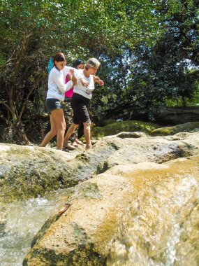 İki genç kadın, Neiva-Huila-Kolombiya 'da bir tatil gezisinde taşlardan bir nehirde yürüyen Latin bir büyükanneye yardım ediyor. Yaşlılar, seyahat ve yardım kavramı