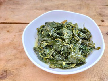 Ahşap bir masada beyaz bir kasede servis edilen manyok yapraklarının yakın görüntüsü. Hindistan cevizi sütü ve baharatıyla pişirilmiş geleneksel bir Endonezya yemeği.. 