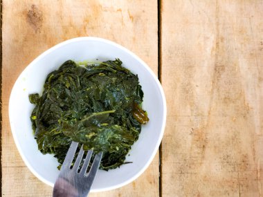 Ahşap bir masada beyaz bir kasede servis edilen manyok yapraklarının yakın görüntüsü. Hindistan cevizi sütü ve baharatıyla pişirilmiş geleneksel bir Endonezya yemeği.. 