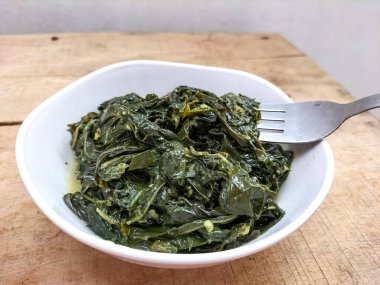 Ahşap bir masada beyaz bir kasede servis edilen manyok yapraklarının yakın görüntüsü. Hindistan cevizi sütü ve baharatıyla pişirilmiş geleneksel bir Endonezya yemeği.. 