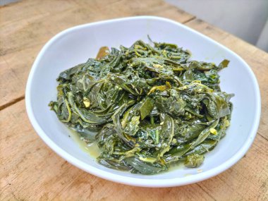 Ahşap bir masada beyaz bir kasede servis edilen manyok yapraklarının yakın görüntüsü. Hindistan cevizi sütü ve baharatıyla pişirilmiş geleneksel bir Endonezya yemeği.. 