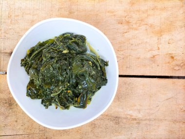 Ahşap bir masada beyaz bir kasede servis edilen manyok yapraklarının yakın görüntüsü. Hindistan cevizi sütü ve baharatıyla pişirilmiş geleneksel bir Endonezya yemeği.. 