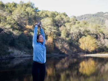 Arka planı olarak sakin bir göl olan bir kadın, kollarını gökyüzüne doğru uzatarak sabah esneme hareketi yapar.