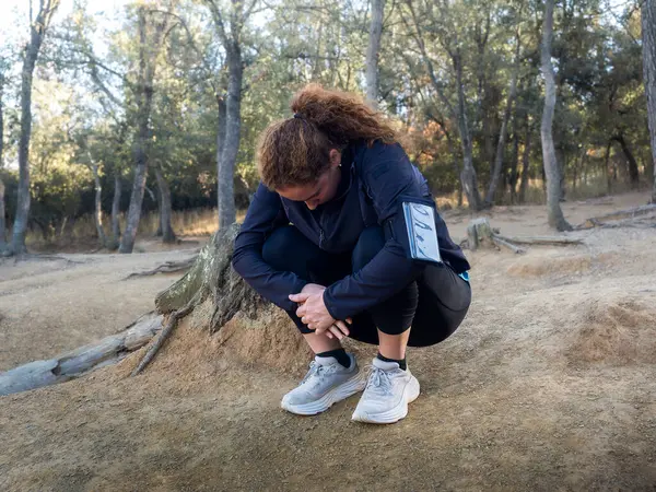 A woman takes a moment to stretch, crouching down on a forest trail, during an outdoor workout session